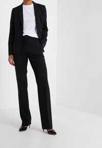 Černý oblek na míru s blejzrem na jeden knoflík, kombinovaný s bílým tričkem. Dlouhé rovné kalhoty, které jsou nošeny s černými botami na jehlovém podpatku.