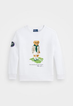 Weißes Sweatshirt mit langen Ärmeln, das einen gestickten Bären in Sportkleidung zeigt, mit einem grünen Schal und einem Tennisschläger vor einem Gras-Hintergrund.