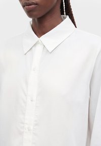 Femme aux cheveux tressés portant une chemise blanche boutonnée avec un col pointu sur un fond uni.