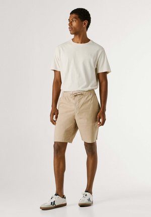 Man die beige korte broek, wit T-shirt en witte sneakers draagt, staat en naar links kijkt tegen een effen achtergrond.