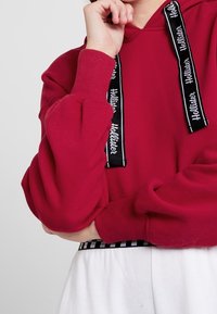 Felpa rossa con cappuccio, maniche lunghe, polsini a coste e lacci neri con testo bianco. Abbinata a pantaloni bianchi e una cintura a scacchi.