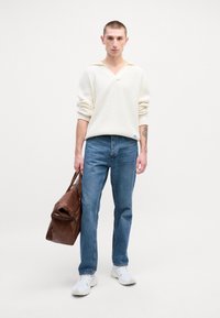Hvit strikket genser med V-hals, blå jeans og hvite sneakers. Holder en brun skinnveske. Enkel, ren design. Ingen mønstre eller aksenter.