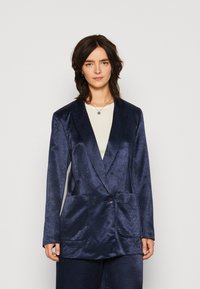 Blazer satinat bleumarin, cu două rânduri de nasturi, două buzunare frontale, guler cu rever și textură netedă, purtat deasupra unei bluze de culoare deschisă.