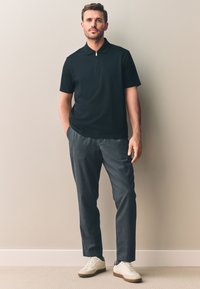 Zwart poloshirt met een kraag met rits, gecombineerd met donkergrijze broek. Gestyled met lichte sneakers, tegen een neutrale achtergrond.