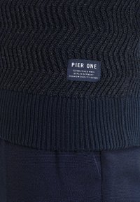 Pull en maille bleu marine avec motif chevron et ourlet côtelé, avec une étiquette Pier One indiquant « Established MMXI Berlin-Germany Premium Quality Goods. »