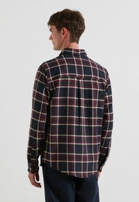 Heren blouse met lange mouwen in een geruit patroon in marineblauw, rood en crème, met een knoopsluiting bij de kraag, een achterpand en een rechte zoom met zijslits.
