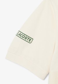 Polo couleur crème en tissu texturé, avec un logo "LACOSTE" brodé en vert sur la manche gauche. Manches courtes, coupe standard.