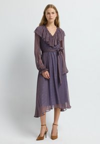 Vloeiende paarse jurk met een ruchesnek, lange mouwen en een strikdetail in de taille. Transparante textuur met een patroon; enkel-lang.
