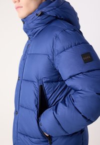 Homme portant une veste matelassée bleue avec capuche, main dans la poche zippée, et un patch logo BOSS noir sur la manche supérieure.