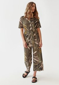 Blomstret jumpsuit i olivengrøn, med korte ærmer, bælte i taljen og brede ben med kontrastfarvede bladmønstre. Sorte slidesandaler.