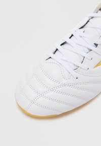 Hvit fotballsko med teksturert lær-overdel, fremtredende sømmer og gul gummisåle. Snøring er synlig på toppen.