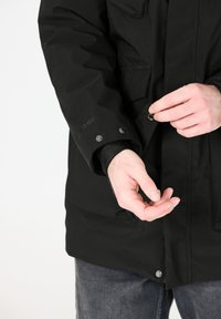 Musta vedenpitävä takki korkealla kauluksella, useilla taskuilla ja napsautettavilla kiinnityksillä. Siinä on logo hihassa ja teksturoitu kangas.