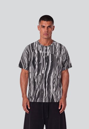Uomo che indossa una camicia a maniche corte nera e grigia con motivo e testo "COLUCCI" davanti e pantaloni neri su uno sfondo grigio uniforme.