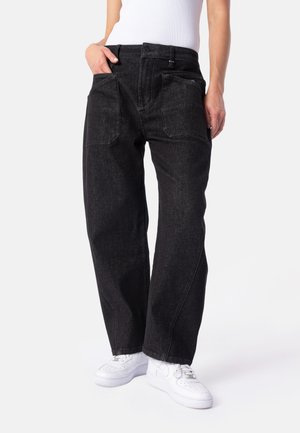 Person trägt lockere schwarze Denim-Jeans mit großen Fronttaschen, ein weißes ärmelloses Oberteil und weiße Sneakers, steht vor einem schlichten Hintergrund.