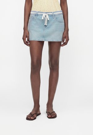 Persoon draagt een lichtblauwe denim minirok met een witte trekkoord, een geel bloementopje en bruine slippers, staand tegen een witte achtergrond.
