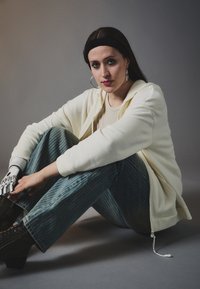 Hvit zip-up hettegenser, beige topp, stripete blå bukser, svarte snøring støvler, og en sølv protesehånd. Ovale øredobber og mørk hårbånd.