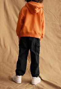 Oranžová mikina s kapucňou, čierne cargo nohavice s vreckami a biele sneakersy. Mäkka látka, voľný strih, elastické manžety na nohaviciach.