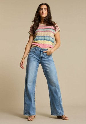 Vrouw staand met hand in de zak, gekleed in een meerkleurige gestreepte top met korte mouwen, wijd uitlopende blauwe spijkerbroek en tankleurige sandalen tegen een beige achtergrond.