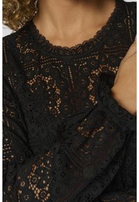 Haut noir en dentelle à manches longues avec des motifs floraux et géométriques porté par une personne aux cheveux bouclés et aux bras croisés.