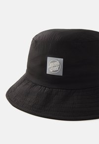 Chapeau seau noir en tissu léger avec un bord rond cousu. Un patch carré avec "SANTA CRUZ" est fixé à l'avant.
