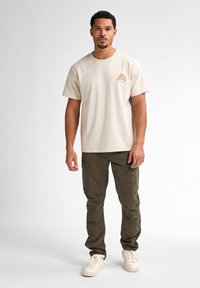 T-shirt en coton beige à manches courtes, arborant un motif marron sur le côté gauche du torse ; associé à un pantalon cargo vert foncé et des baskets blanches.