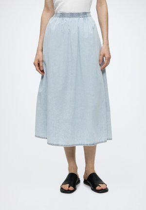 Femme portant une jupe midi bleu clair avec taille élastique, haut blanc et sandales noires à enfiler, debout devant un fond uni.