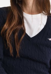 Pull en tricot bleu marine avec un motif torsadé et un col en V, superposé à une chemise blanche. Comprend un petit logo sur la poitrine.