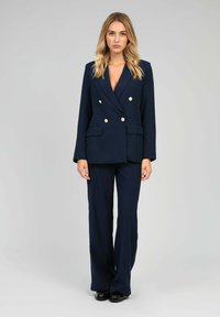 Costume ajusté bleu marine avec un blazer croisé à double boutonnage et boutons dorés, pantalon à jambes larges et un tissu lisse et structuré.