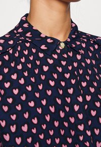 Blusa blu navy con motivo a cuori rosa, con colletto e chiusura a bottoni dorati. Tessuto liscio e leggero.