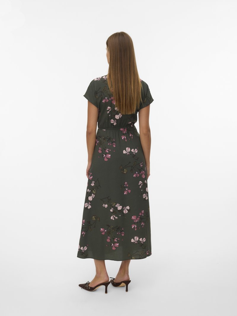 Robe maxi florale verte à manches courtes, ornée de motifs de fleurs roses et blanches, taille cintrée et tissu fluide. Sandales à talons brunes.