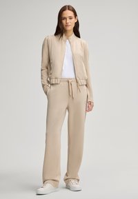 Giacca bomber beige con zip, t-shirt bianca sotto e pantaloni jogger beige coordinati. Tessuto strutturato, vita elasticizzata e tasca.