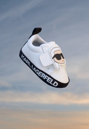 Basket blanche pour tout-petit avec semelle noire, marquée "Karl Lagerfeld" et visage de dessin animé avec des lunettes de soleil sur la sangle Velcro, sur fond de ciel bleu.