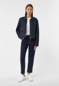Giacca di denim scuro con due tasche frontali, cuciture bianche in contrasto, abbinata a pantaloni di denim scuro e sneakers bianche.