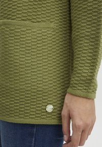 Pull en tricot texturé vert olive avec une poche avant et un accent de bouton de couleur claire. Manches longues et poignets côtelés.