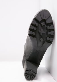 Bota de cuero negra con suela texturizada, que presenta profundas estrías y un tacón cuadrado, apoyada en un fondo blanco.
