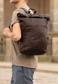 Smeđa kožna ruksak s preklopom, teksturirane površine, s dekorativnim šavovima i gornjom drškom. Podesive naramenice, minimalistički dizajn.