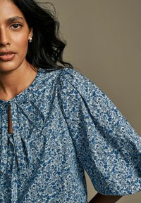 Cvetna bluzica z modro-belo vzorcem, okroglo izrezana, z detajlom v obliki ključavnice in obsežnimi pufastimi rokavi. Lahka tkanina.