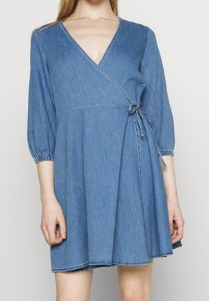 Kvinde iført en blå denim slå-om-kjole med trekvartlange pufærmer og lukning med bindebånd i siden.