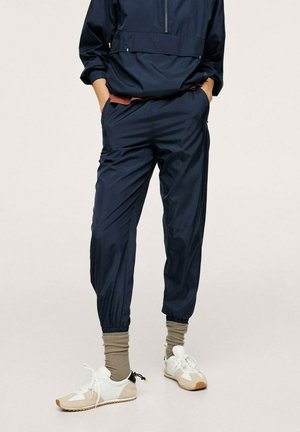 Personne portant un pantalon de jogging bleu marine, des chaussettes hautes beige et des baskets blanches et beiges, debout les mains dans les poches contre un fond uni.