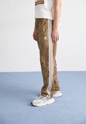 Person trägt braune Adidas-Trainingshose mit Muster, weißen Streifen und silbernen Adidas-Sneakers, steht auf blauem Boden vor weißer Wand.