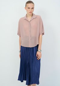 Blusa a maniche corte di un leggero rosa pallido con bottoni sul davanti, abbinata a una gonna blu navy fluida e plissettata. Tessuto morbido e vestibilità rilassata.