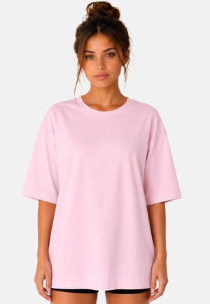 Jeune femme aux cheveux bruns en chignon, portant un t-shirt ample rose clair à manches courtes et un short noir sur un fond uni.