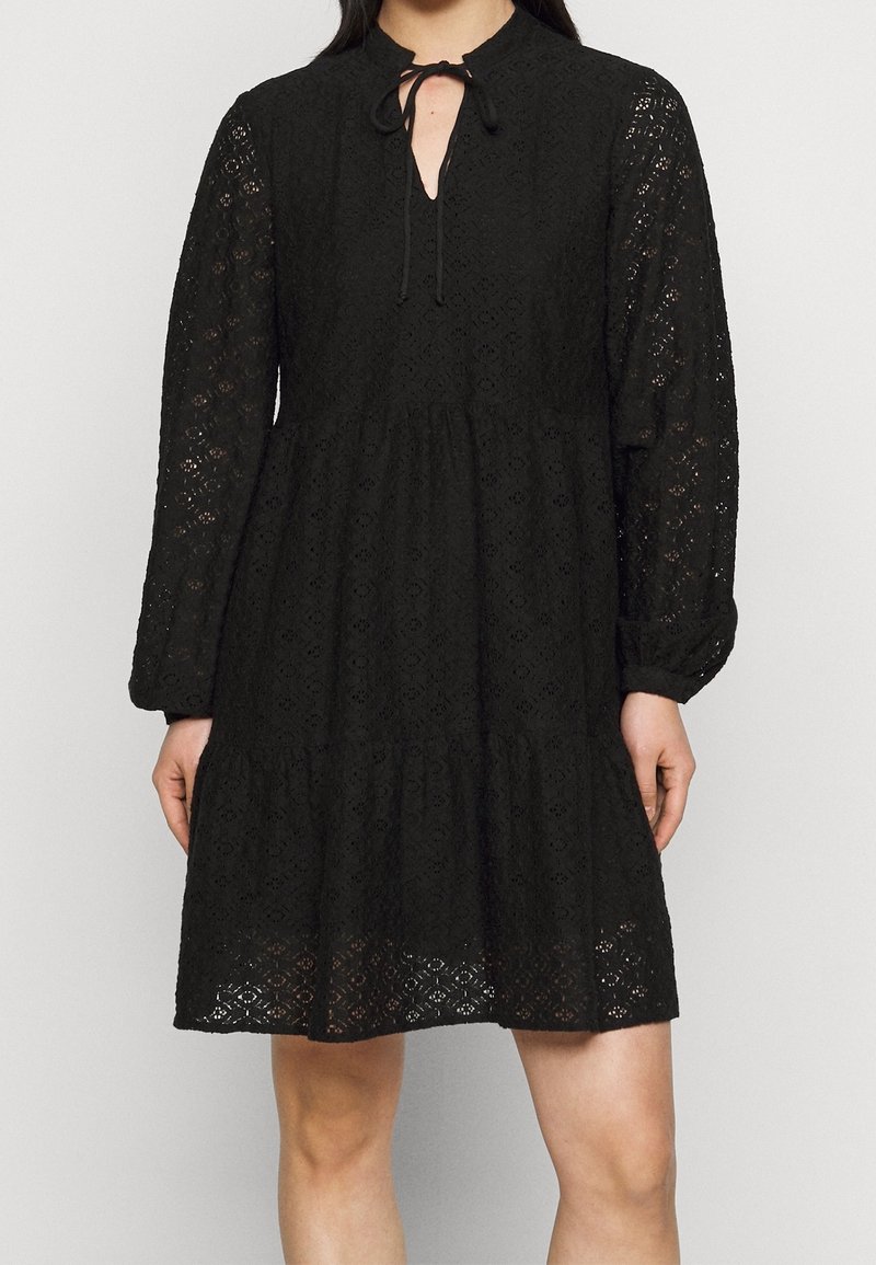 Robe en dentelle noire longueur genou avec manches longues bouffantes et un petit nœud au col, portée par une personne debout.