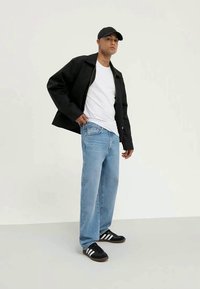 Svart hettejakke med glidelås, hvit t-skjorte, lyseblå jeans og svarte joggesko med hvite striper. Modell med svart caps står foran en ensfarget bakgrunn.
