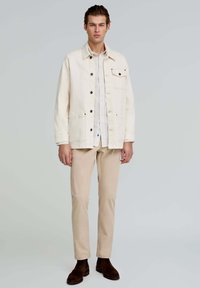 Chaqueta de mezclilla beige claro con bolsillos frontales y botones negros, superpuesta a una camisa blanca a rayas, combinada con pantalones beige y botas marrones.