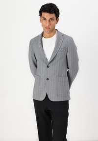 Blazer a righe grigie con rever a punta, due tasche frontali e due bottoni. Indossato sopra una camicia bianca, abbinato a pantaloni neri.
