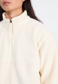 Kermanvärinen fleece-pullover, jossa on puoli-vetoketju edessä, korkea kaulus ja brodeerattu logo. Pehmeä teksturoitu kangas, jossa näkyvät ommeltyöt.