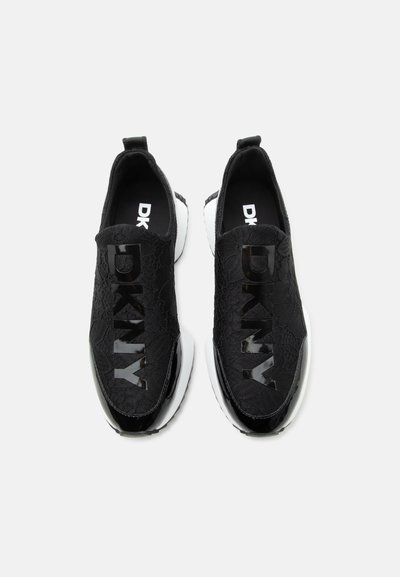 Baskets noires à enfiler avec semelles blanches, dessus texturé et grand logo brillant DKNY sur le dessus de chaque chaussure.