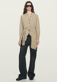 Vrouw draagt een beige vest met knopen en een bijpassende trui om haar middel gestrikt, zwarte wijde broek, zwarte schoenen en een zonnebril.