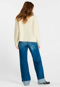 Vrouw met lang bruin haar, gekleed in een crèmekleurige fuzzy trui, wijde blauwe geopperde jeans en zwarte schoenen, staand met de rug naar ons toe.